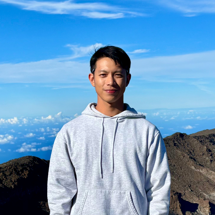 C. J. Huang standing outdoors on a mountain summit in Hawaii, wearing a light gray hoodie with blue sky and ocean in the background.